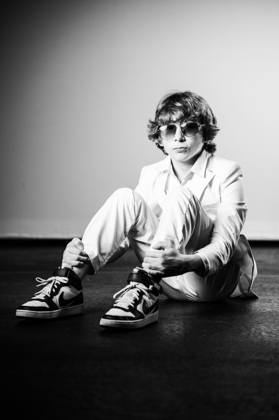 Caelum, founder of Battery Powered Bird, sitting on a studio floor in a white suit and sunglasses
